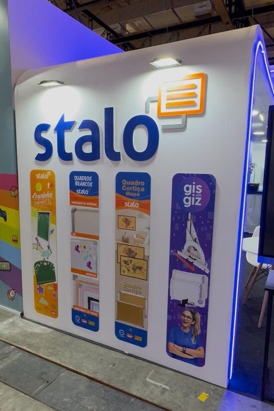 Estande da marca "Stalo" com quatro banners verticais coloridos, cada um destacando uma linha de produtos: Infantil (laranja), Quadros Brancos (azul claro), Quadro Cortiça (amarelo) e Gis com Giz (roxo), com imagens ilustrativas.
