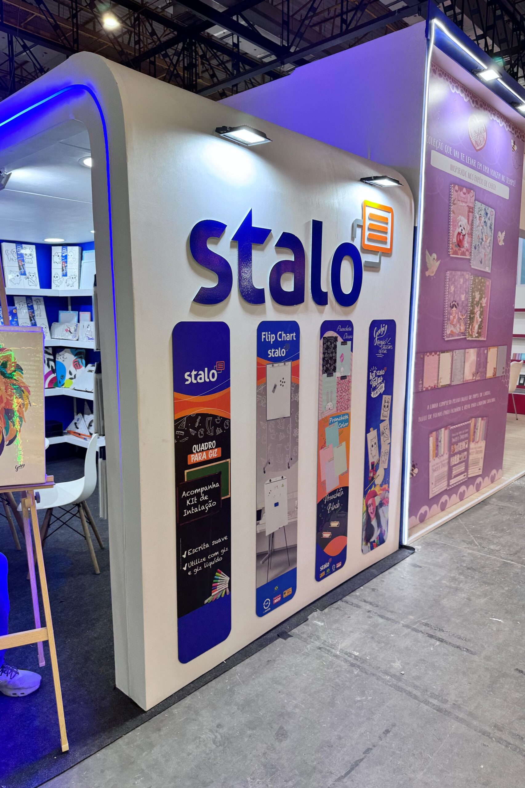 Estande da marca "Stalo" com estrutura branca e iluminação superior. Na parede frontal, há imagens coloridas de produtos como flip charts e materiais de papelaria. À direita, um painel roxo exibe cadernos decorados. Dentro do estande, há prateleiras com itens e uma mesa com cadeira branca.