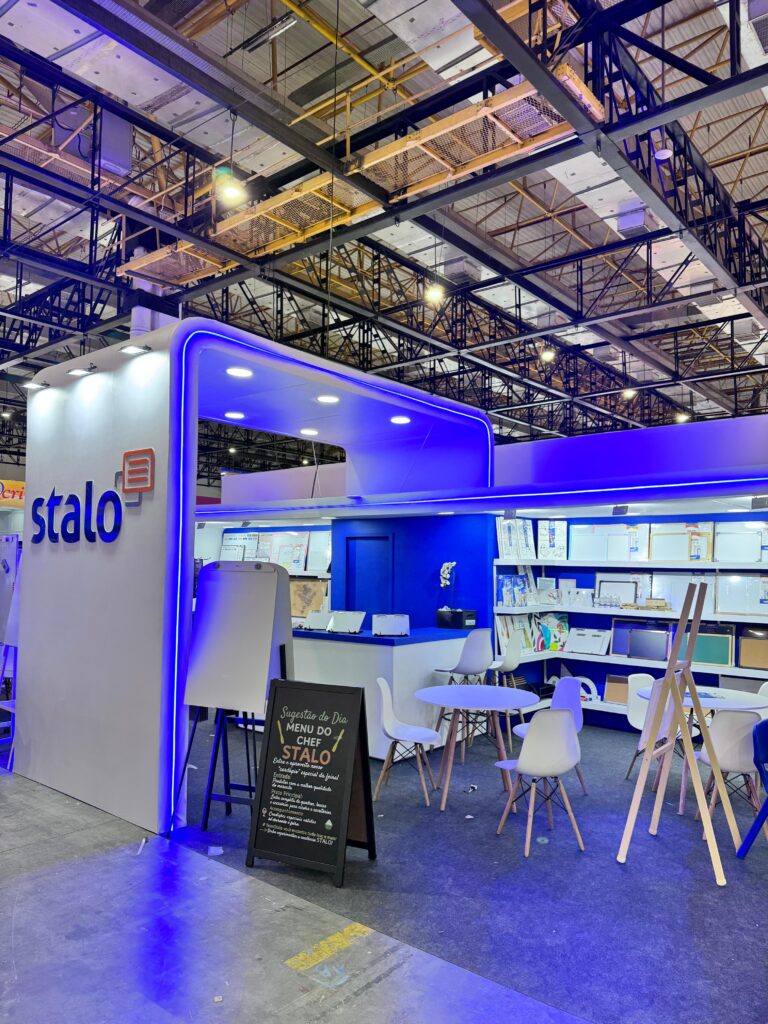 Estande da marca "Stalo" com iluminação azul, parede branca com logotipo, mesa redonda com cadeiras brancas e quadro negro com texto em giz. Ao fundo, prateleiras exibem quadros brancos e materiais diversos.