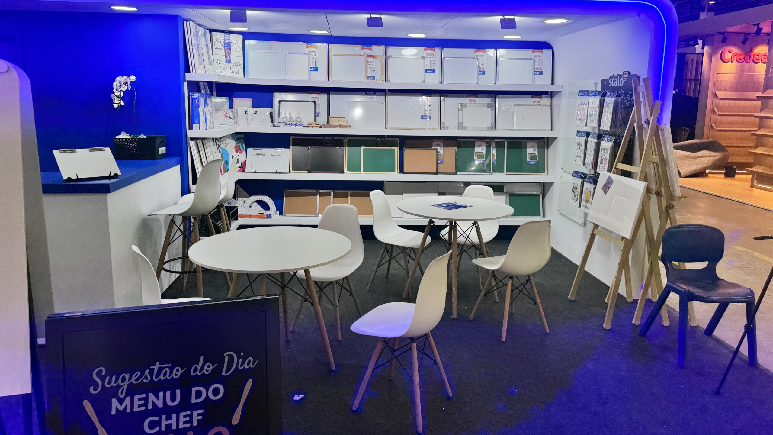 Área de exposição com mesas redondas brancas, cadeiras pretas e brancas, e prateleiras ao fundo com quadros brancos e materiais de escritório. À esquerda, há um balcão com vaso de flores e um quadro negro com a frase "Sugestão do Dia MENU DO CHEF". À direita, cavaletes exibem mais produtos.