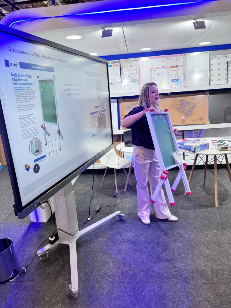 Mulher segura uma pequena lousa verde com pernas brancas e detalhes vermelhos, enquanto observa uma tela interativa com instruções de montagem. A sala tem cadeiras, mesas e mapas nas paredes ao fundo.