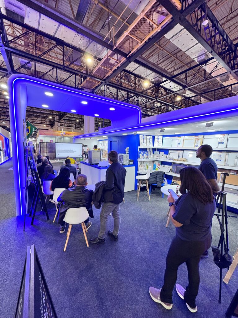 Estande de exposição com iluminação azul, onde pessoas estão sentadas em cadeiras brancas ou em pé. Ao fundo, há prateleiras com produtos e uma tela grande usada para apresentação.