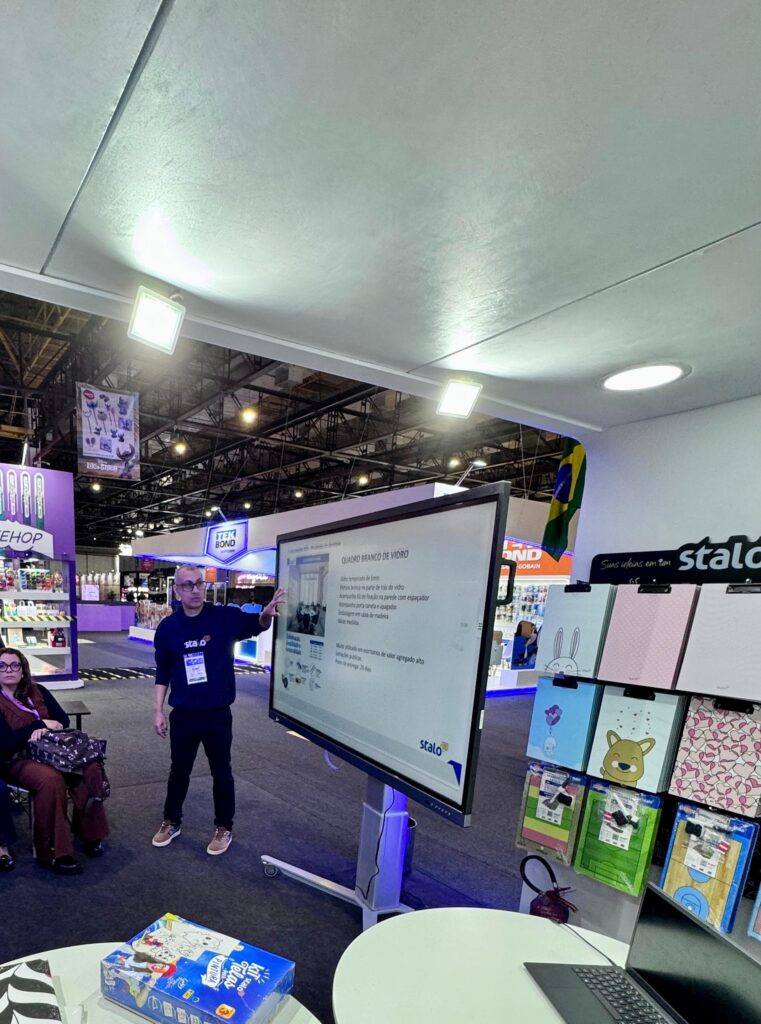 Pessoa apresentando em uma tela grande durante uma feira ou exposição. A tela exibe um slide com texto sobre o produto Quadro Branco de Vidro, destacando sua versatilidade, desempenho e interface fácil de usar. No canto inferior direito da tela está o logotipo 'Stalo'. Ao fundo, há estandes de empresas com produtos expostos e, no canto inferior esquerdo da imagem, uma mesa redonda branca com alguns itens sobre ela.