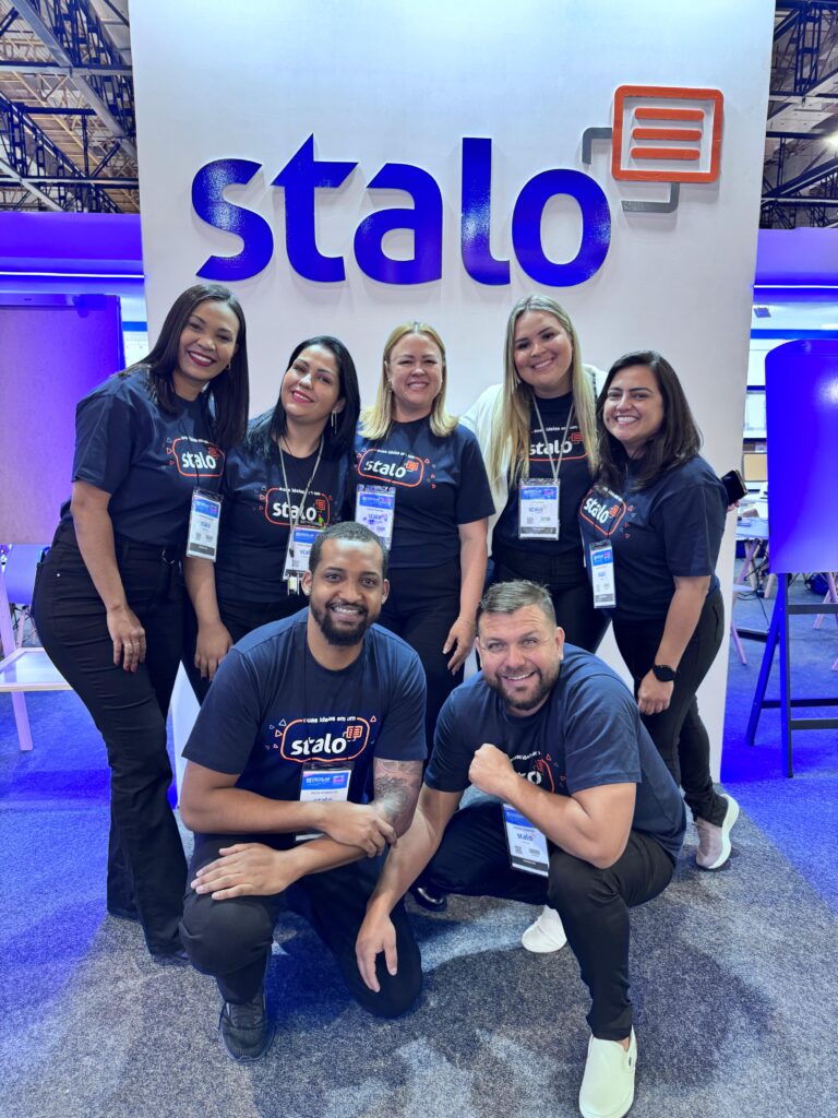 Grupo de sete pessoas posando para foto em frente a um estande com a palavra 'stalo' em destaque. Todos usam camisetas azul-escuras com o logotipo 'stalo' e crachás pendurados no pescoço. Ao fundo, há uma placa iluminada e elementos de exposição, indicando que a imagem foi tirada em um evento ou conferência.