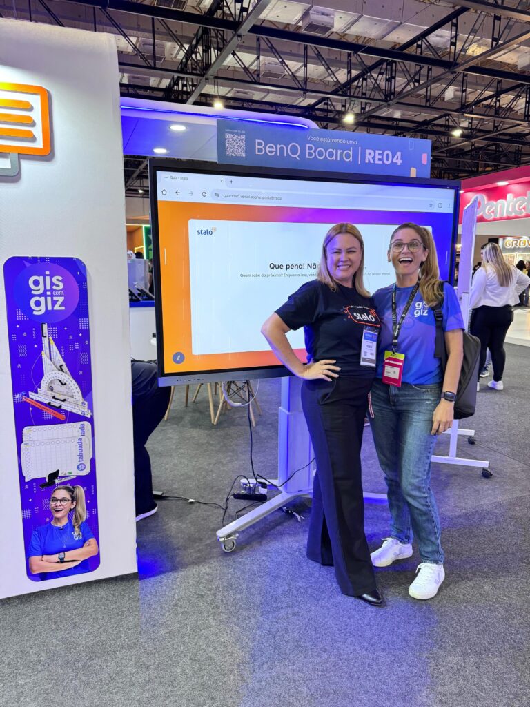 Duas pessoas em pé posam para foto em frente a um quadro interativo BenQ Board | RE04. À esquerda, há um banner vertical com o texto 'gis giz' e imagens de materiais escolares. O ambiente é uma feira ou exposição, com estandes e outras pessoas ao fundo.