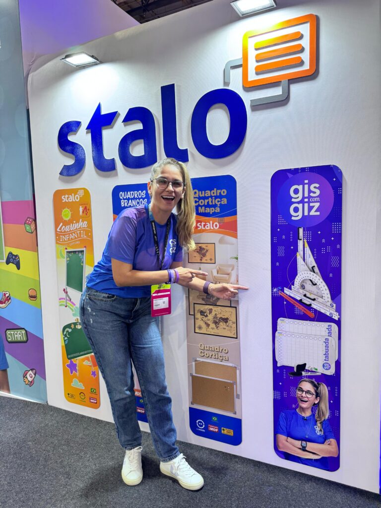 Pessoa em pé apontando para um display de produtos da marca 'stalo', que inclui quadros brancos e quadros de cortiça. Ao lado, há uma seção com o título 'gis com giz', exibindo imagens de giz e um quadro branco. A pessoa veste camiseta azul, jeans, tênis brancos e crachá pendurado no pescoço.