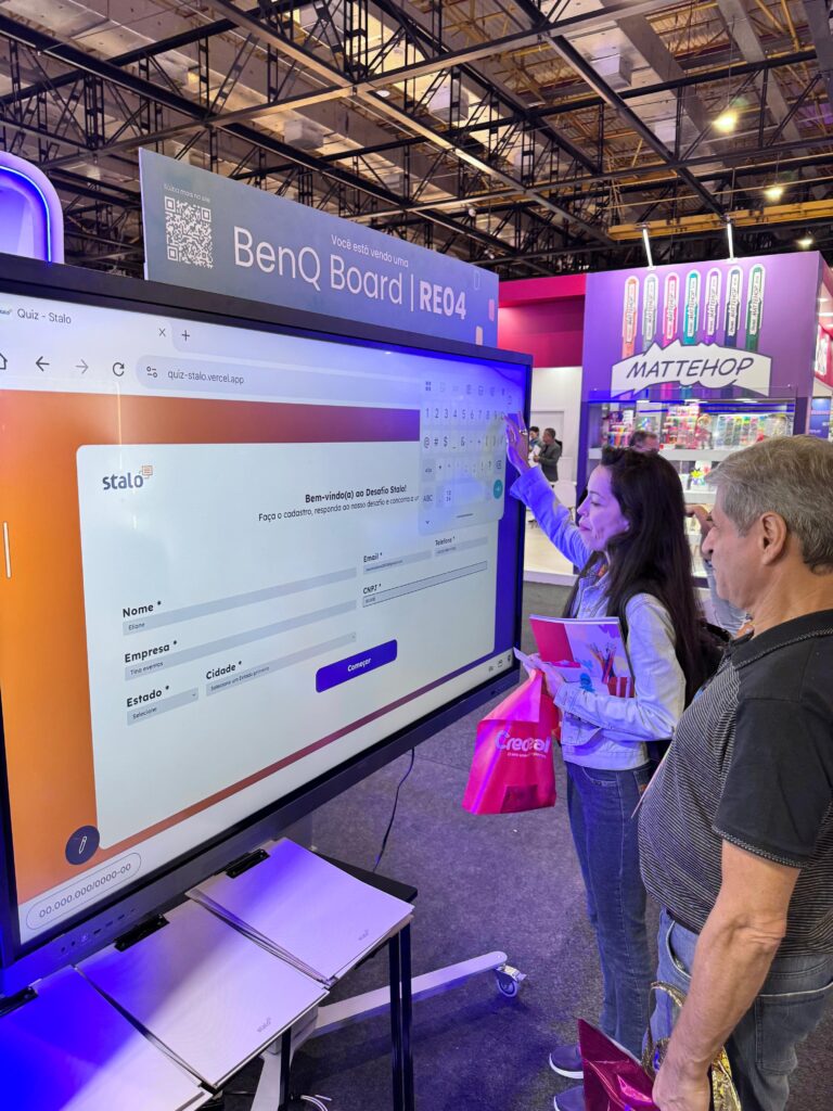 Duas pessoas em frente a um quadro interativo BenQ Board | RE04, em uma feira ou evento. Uma delas toca a tela, que exibe uma interface de formulário com campos para 'Nome', 'Empresa' e 'E-mail'. Ao fundo, há estandes de outras empresas.
