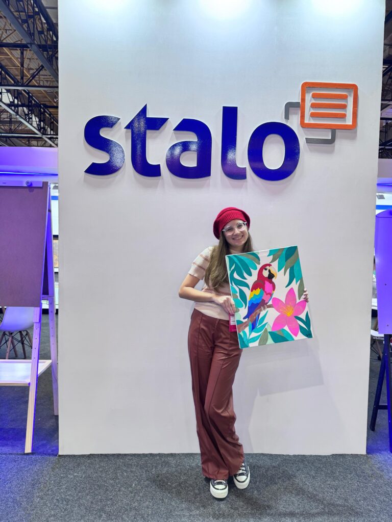 Pessoa em pé em frente a uma parede branca com a palavra 'stalo' escrita em letras azuis. Ela segura uma pintura colorida de um papagaio cercado por flores e folhas. A pessoa veste chapéu vermelho, camisa branca e calça vinho. Há cadeiras ao lado e elementos de iluminação roxa ao fundo.