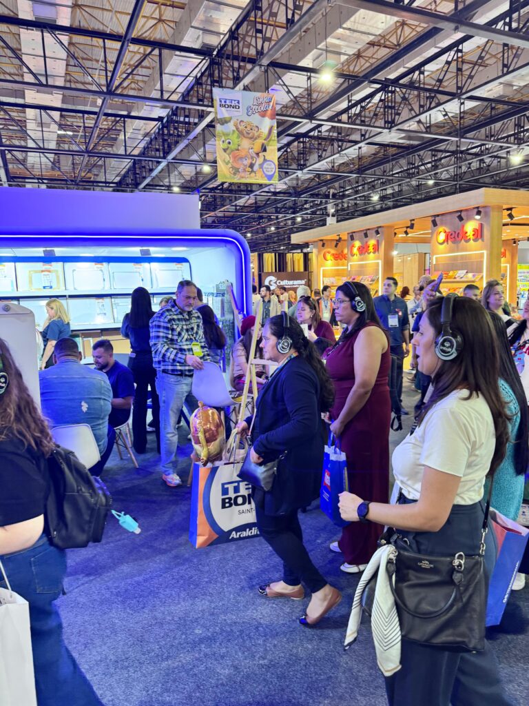 Grupo de pessoas em uma feira ou evento, com algumas em pé e outras sentadas. Ao fundo, há um estande iluminado com luzes azuis, onde pessoas estão sendo atendidas. O teto possui estrutura metálica com iluminação. Outros estandes com letreiros luminosos são visíveis.