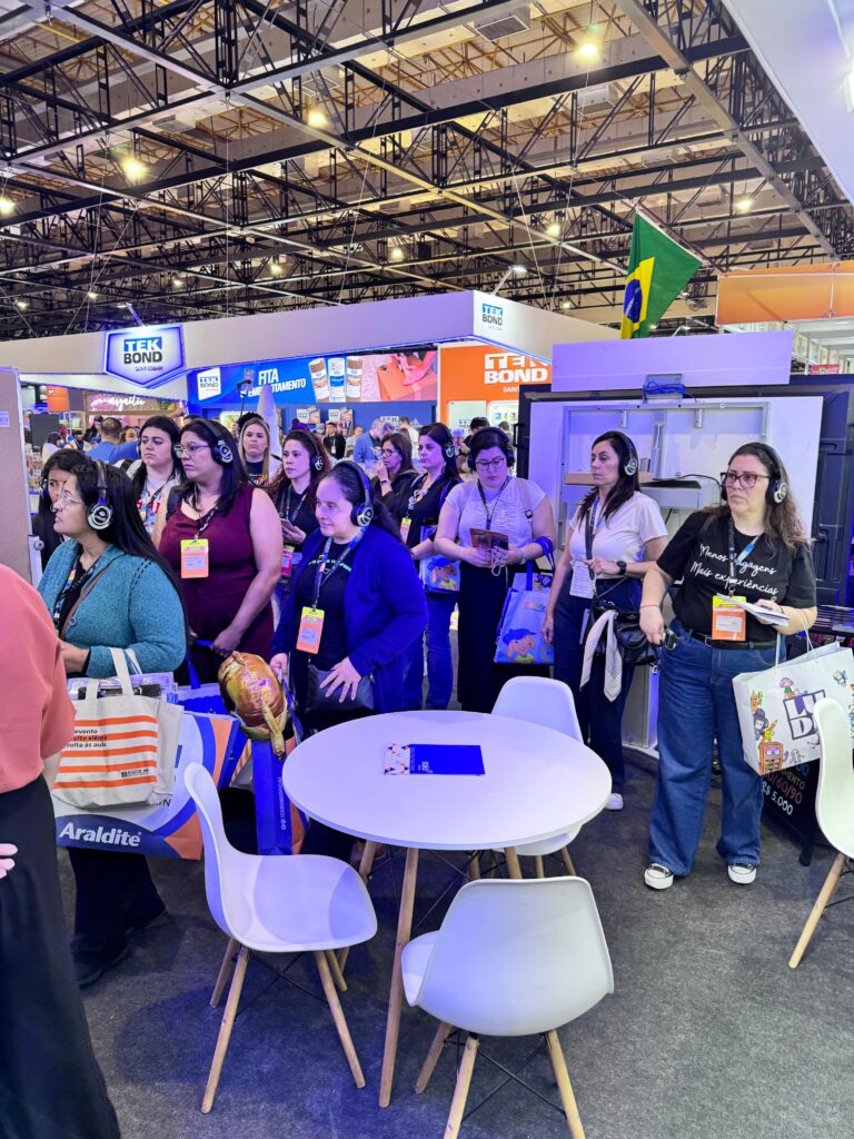 Grupo de pessoas em pé ao redor de uma mesa redonda branca com quatro cadeiras, em um evento ou feira. Todos usam fones de ouvido e carregam sacolas, alguns com crachás pendurados no pescoço. Ao fundo, há estandes com banners e produtos expostos, e uma bandeira do Brasil visível no canto superior direito.