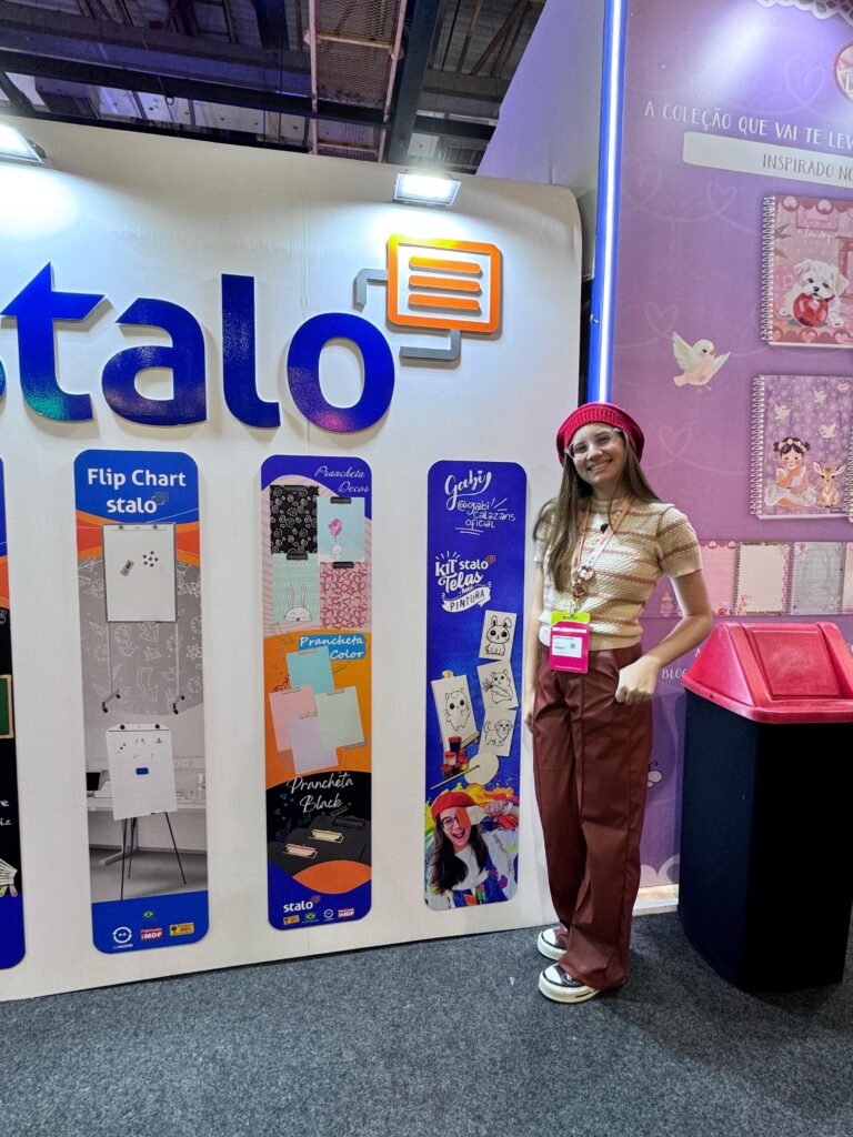 Mulher posa ao lado de estande da marca "Stalo", com logotipo azul e quatro banners verticais: Flip Chart, Pranchetas, trlas de pintura. Ela veste camiseta listrada, calça marrom larga, boné rosa e crachá. Ao fundo, há imagens infantis e um cesto rosa.