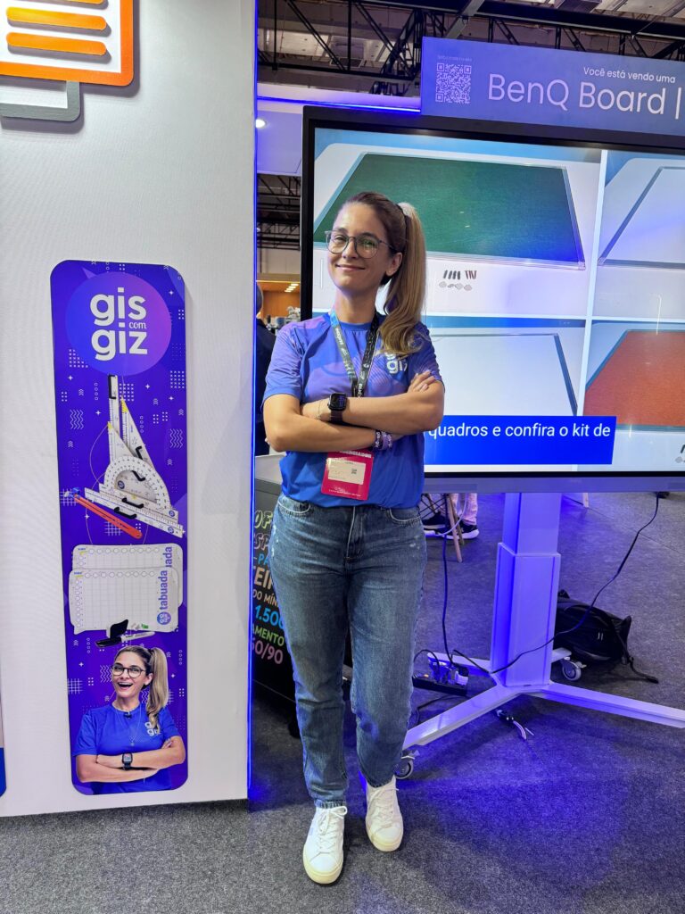 Mulher de braços cruzados em frente a um monitor grande que exibe imagens de quadros brancos. Ela veste camiseta azul com o logo "giz com giz" e crachá. Ao lado, há um banner roxo com o mesmo logo e imagem da mulher.