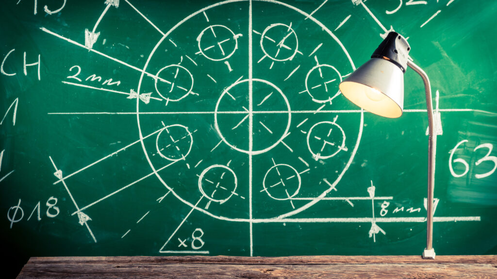 Um quadro-negro verde com um desenho técnico detalhado em giz branco de um objeto circular com medidas. Uma luminária de mesa cinza acesa está sobre uma mesa de madeira rústica em frente ao quadro, iluminando a cena.